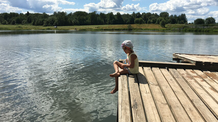 funny country alone barefoot child in cap sits on the edge of a wooden bridge over the water and...