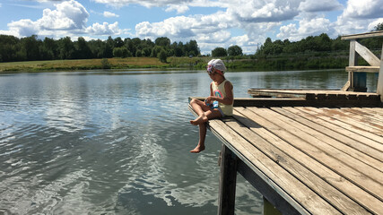 funny country alone barefoot child in cap sits on the edge of a wooden bridge over the water and...