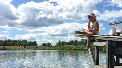 funny country alone barefoot child in cap sits on the edge of a wooden bridge over the water and...