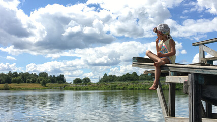 funny country alone barefoot child in cap sits on the edge of a wooden bridge over the water and look around. natural background sky with clouds - obrazy, fototapety, plakaty