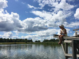 Fototapeta premium country alone barefoot child in cap sits on the edge of a wooden bridge over the water and watch. natural background sky with clouds