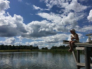 country alone barefoot child sits on the edge of a wooden bridge over the water and watch. natural background sky with clouds