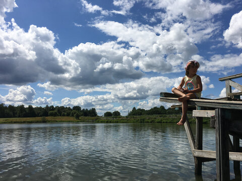 funny country alone barefoot child in cap sits on the edge of a wooden bridge over the water and look around. natural background sky with clouds