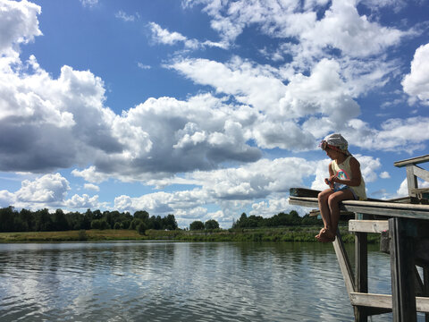country alone barefoot child in cap sits on the edge of a wooden bridge over the water and watch. natural background sky with clouds