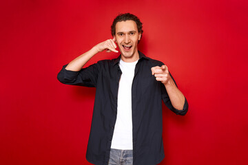 call me hand sign. portrait of young smiling man pointing index finger in front of him depicts telephone receiver in his hand. isolated on red studio background. concept - love, romance, communication