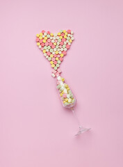 Multicolored marshmallows pour out of a tall glass in the shape of a heart on a pink background. The concept of Valentine's Day.