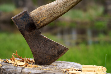 Old rusty axe with wooden handle stuck in the stump. blurred background with pile of wood logs,...