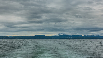 On the surface of the Pacific Ocean, ripples are visible, a foam trail from the yacht. A mountain range against a cloudy sky in the distance. A snow-covered volcano is hiding in the clouds. Kamchatka