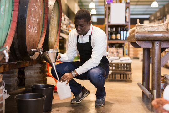 African American seller pouring draft wine from wooden barrels to small plastic can in local shop