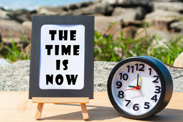 Motivational quote on standing notepad with black alarm clock. The time is now
