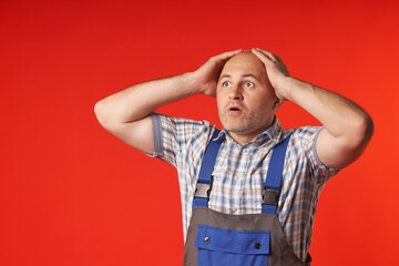 Emotional portrait of a man with his mouth open in overalls, holding his bald head with his hands and looking up against a red background.