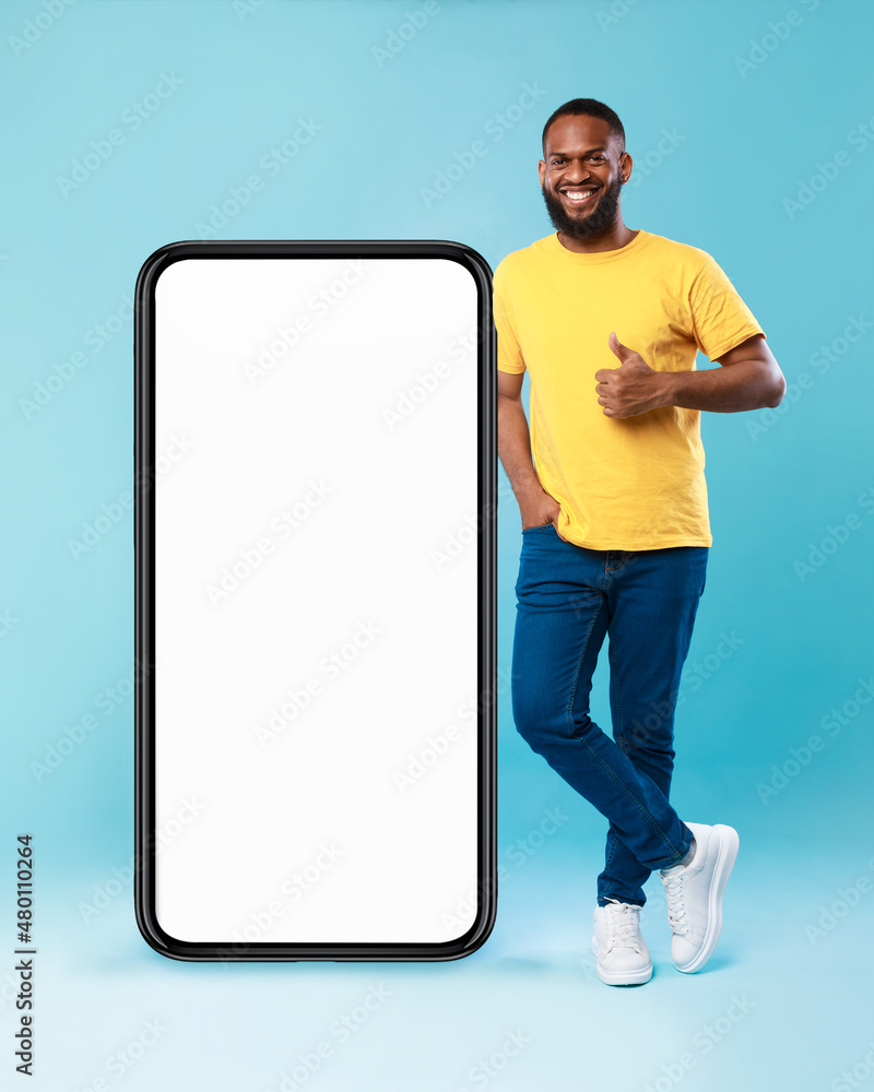 Poster joyful afro guy in gesturing thumb up near giant smartphone over blue background, mockup for website