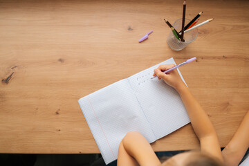 High-angle view of unrecognizable elementary little child girl doing homework writing in copybook with pen sitting at table. Close-up cropped shot of primary little school girl learning at home. - obrazy, fototapety, plakaty