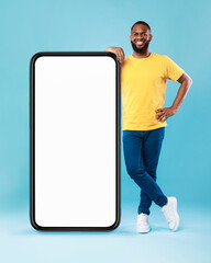 Cheerful Afro guy leaning on giant smartphone, demonstrating empty screen, space for online ad, mockup for mobile app