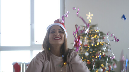 Young beautiful happy woman in deer antlers blows up a petard on the background of a Christmas tree