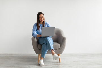 Happy Caucasian lady sitting in armchair with modern laptop, studying remotely or having business meeting