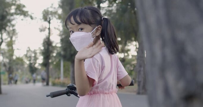 4k Slow motion little Asian girl (5 years old) in a pink dress wearing mask to prevent  spread of coronavirus 19 waving goodbye in the park in the evening and riding bicycle