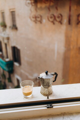old steel Italian geyser coffee maker on a marble windowsill with a cup of hot espresso with milk. coffee prepared in a geyser coffee maker is cooled in the window. morning coffee routine in italy