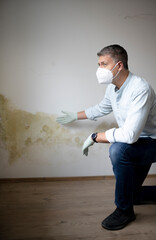 Man with mouth nose mask and blue shirt and gloves n front of white wall with mold