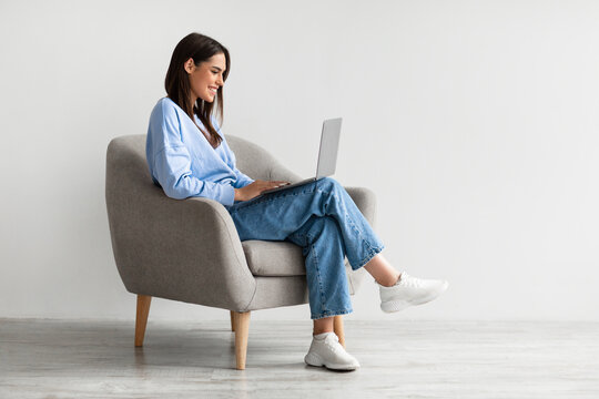 Smiling young woman in casual wear working online, sitting in armchair and using laptop against white wall