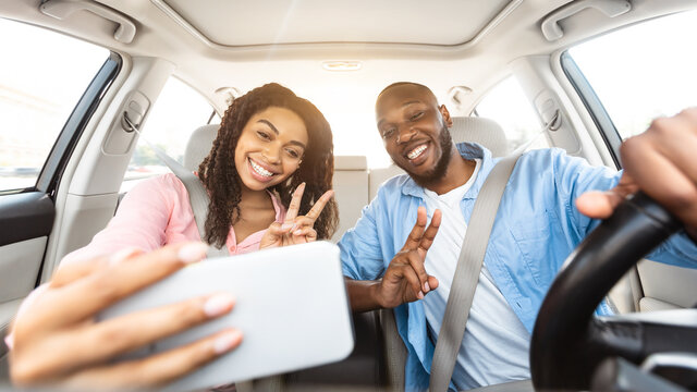 Happy black couple driving car and taking selfie on smartphone