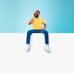 Happy black man sitting on white blank placard, shouting, announcing huge sale on blue background,...