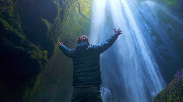 Back View Of A Tourist Man Hiker Raising Arms Wide Admiring The View Facing The Hidden Waterfall Of Gljufrabui In South Iceland. - Low-Angle Shot, Slow Motion