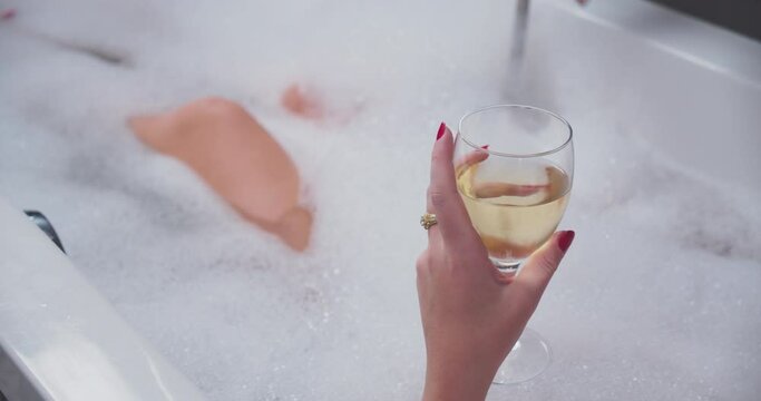 closeup young caucasian girl in bubble bath drinking white wine. Woman leg knee hand with glass.
