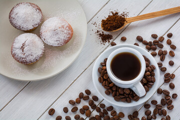 Cupcakes sprinkled with powdered sugar on a plate and a cup of coffee with coffee beans on a white wooden table