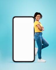 Black woman leaning on cellphone with space for mobile app on white screen, blue background. Smartphone display mockup