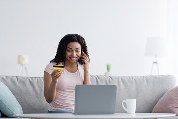 Smiling millennial black woman looks at laptop, hold phone, banking credit card