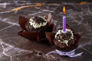 Chocolate cupcakes with white cream and crumbs and burning violet candle in brown paper form on dark marble table.
