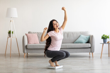 Overjoyed Indian woman sitting on scales, gesturing YES, excited over result of her weight loss...