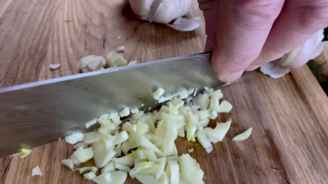 Chef hands chop finely raw garlic on wooden cutting board with kitchen knife. Slow motion.