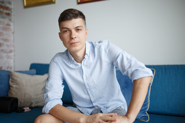 Obraz premium a man of European appearance sits on a blue sofa in his room in a blue shirt and looks thoughtfully at you.