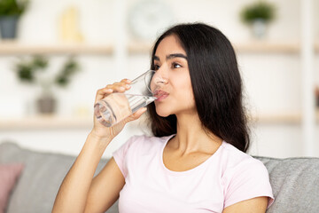 Keep hydrated concept. Attractive young Indian woman drinking water from glass on couch in living...