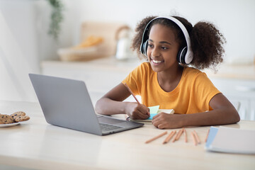 Cheerful black girl studying online, using laptop and headset, drawing