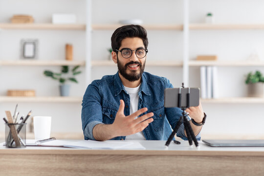 Arab male blogger recording video at home with smartphone on tripod, making content for his blog, sitting at home office