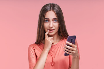 Attractive young long haired caucasian woman holds mobile phone in hand and puts finger in mouth, thinking about what to write to boyfriend flirting by SMS in messenger stands in pink studio.