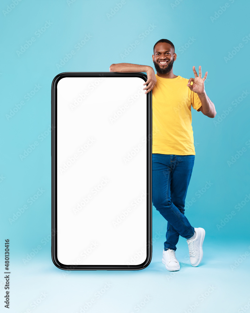 Poster cool black guy showing okay gesture next to huge smartphone on blue background, mockup for mobile ap