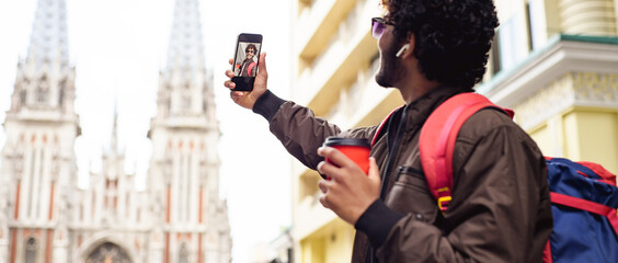 Indian tourist with coffee to go taking selfie on smartphone on urban street, banner shot