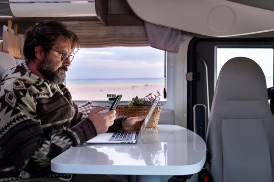Adult man working with laptop inside a camper van. Freedom remote worker lifestyle. Smart working with phone and roaming connection. Camping car interior and beach sunny  day outside the window 