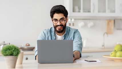 Remote business. Happy arab freelancer man working with laptop in kitchen, typing on computer, enjoying online job