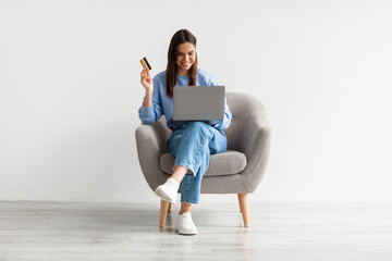 Online shopping, e-commerce, remote banking. Young woman sitting in armchair with laptop and credit...