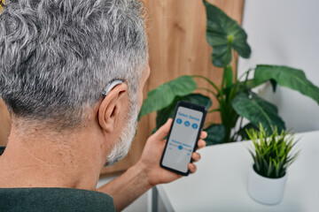 Hearing impaired mature man adjusts settings for his BTE hearing aid via smartphone. Hearing aids, deafness treatment, innovative technologies at audiology - 480136017