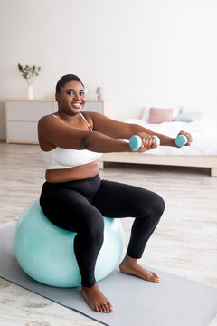 Cheerful Plus Size Afro Woman Exercising With Dumbbells On Fitness Ball At Home, Full Length