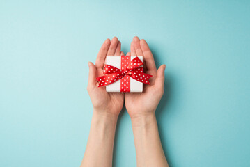 First person top view photo of valentine's day decorations girl's hands holding small white giftbox with red ribbon bow on palms on isolated pastel blue background with copyspace