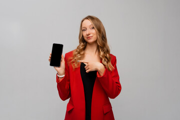 Naklejka premium Business and technology. Beautiful and confident business woman pointing finger at smartphone screen, showing app interface, standing in red blazer against gray background