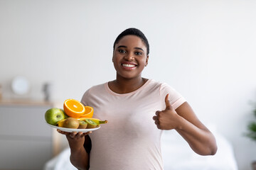 Happy plus size black woman holding fruits and showing thumb up, recommending healthy diet for...