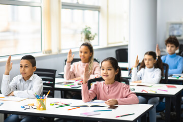 Diverse group of little schoolchildren raising hands at classroom